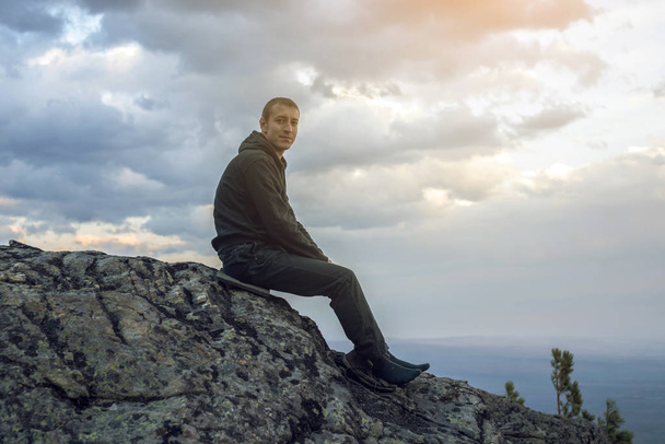 Homem está sentado em uma colina de pedra, mãos olhando para a vista das alturas e goza da liberdade
 - Foto, Imagem