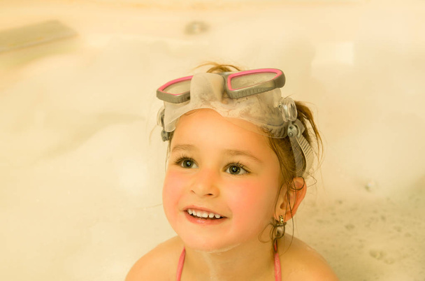 Closeup of a little beautiful girl playing with water and foam in bath using a swimsuit and beautiful dive sunglasses - Foto, imagen