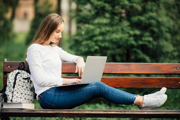 Belle jeune femme allongée sur un banc et travaillant sur un ordinateur portable à l'extérieur
. - Photo, image