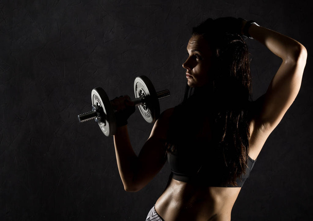 Fitness sexy girl with dumbbells on a dark background. Athlete doing exercises in the gym - Foto, Imagem