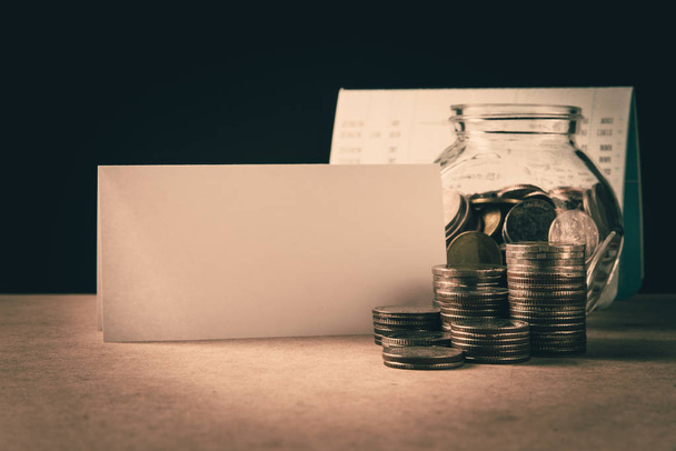 ряд монет и монет в бутылке перед книгой счетов с белой пустой бумагой на деревянном столе и черным фоном в финансово-банковской концепции
 - Фото, изображение