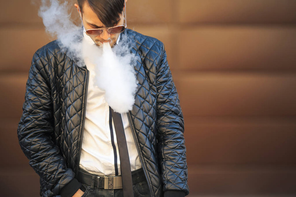 Vape man. Portrait of a handsome young white guy with modern haircut in aviator sunglasses vaping an electronic cigarette opposite the futuristic urban background. - Photo, image
