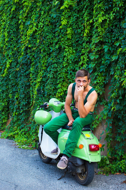 The guy is sitting on a parked scooter against a wall of ivy - Fotoğraf, Görsel