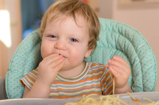 Χαριτωμένο μωρό ενός έτους τρώει μακαρόνια στην παιδική καρέκλα.  - Φωτογραφία, εικόνα