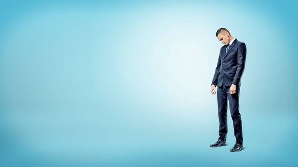 A sad businessman standing on blue background looking down with slumped shoulders. - Фото, изображение