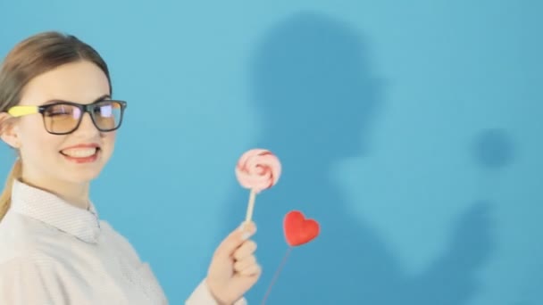 Portrait of Funny Girl with Ponytail and Glasses Holding Heart and Lollipop in Hands Posing in Studio on Blue Background. Cute Brunette with Red Lips in Shirt. - Imágenes, Vídeo