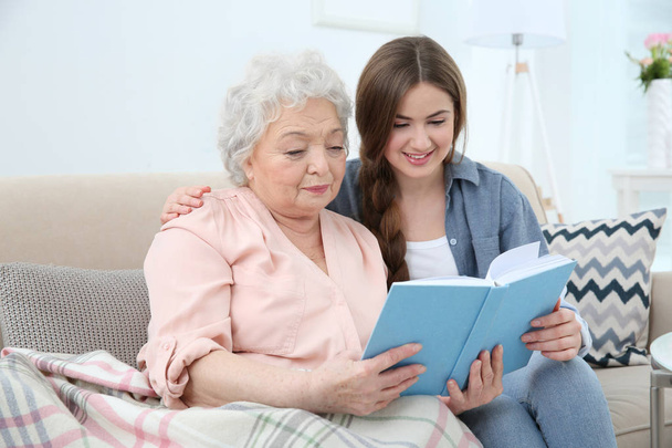 Beautiful girl with grandmother reading book on couch at home - Foto, imagen
