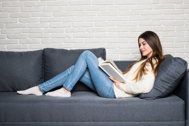 Evdeki kanepede kitap okuyan mutlu genç bir kadın. - Fotoğraf, Görsel
