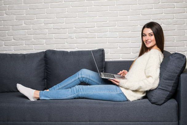 Mulher bonita feliz usando laptop sentado no sofá acolhedor em casa
 - Foto, Imagem