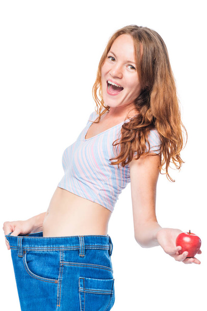 Эмоциональная счастливая девушка после яблочной диеты поддерживает брюки на белой ба
 - Фото, изображение