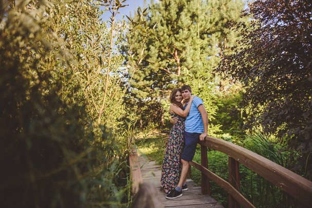 Happy romantic couple in the village, stroll on the wooden bridge. Young beautiful woman in long summer dress hugging the man. Look at the camera. - 写真・画像