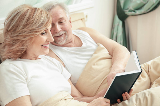 Elderly couple reading book while resting in bed at home - Photo, image