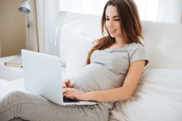 Cheerful pregnant young woman lying and using laptop on bed - Фото, изображение