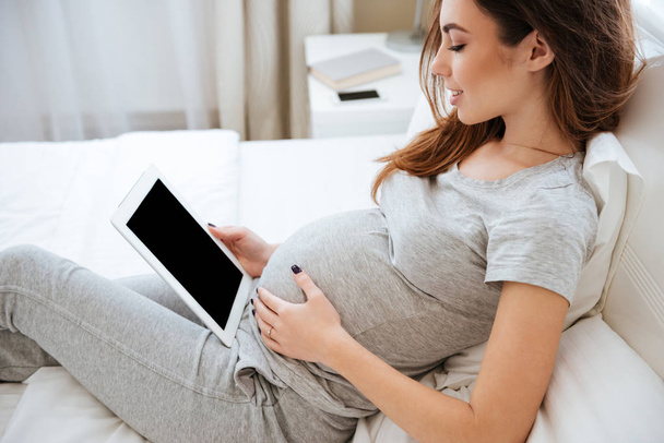 Smiling pregnant young woman lying and using blank screen tablet - Fotó, kép