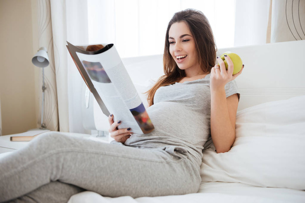 Cheerful pregnant woman eating apple and reading magazine on bed - Photo, Image