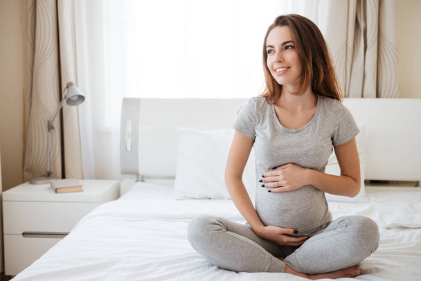 Happy pregnant young woman sitting on bed in bedroom - Foto, afbeelding