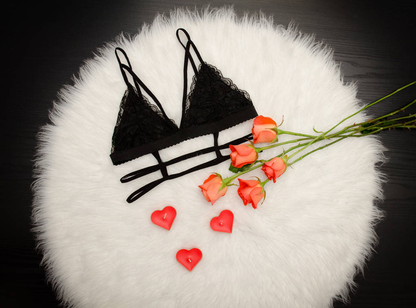 Black lace bra on white fur and a bouquet of orange roses. Fashionable concept. Top view - Photo, Image