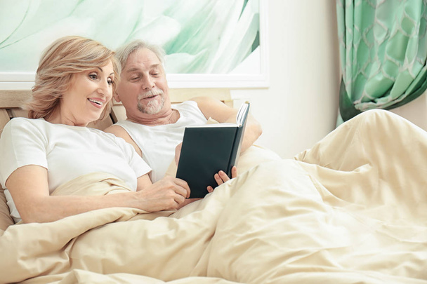 Elderly couple reading book while resting in bed at home - Фото, изображение