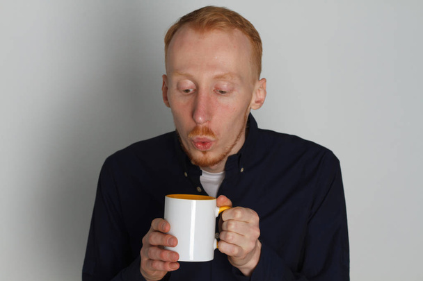 A young man with a mug of tea or coffee. He pleased. White background. Redhead male with white mug. - Фото, зображення