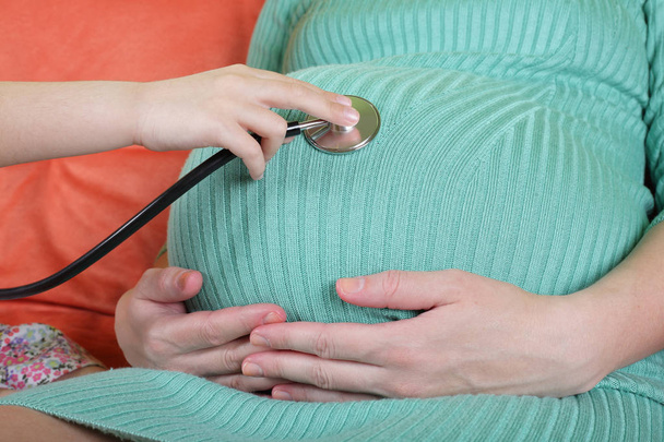 Tytär käyttäen stetoskooppi valkoinen tutkii raskaana äiti
 - Valokuva, kuva