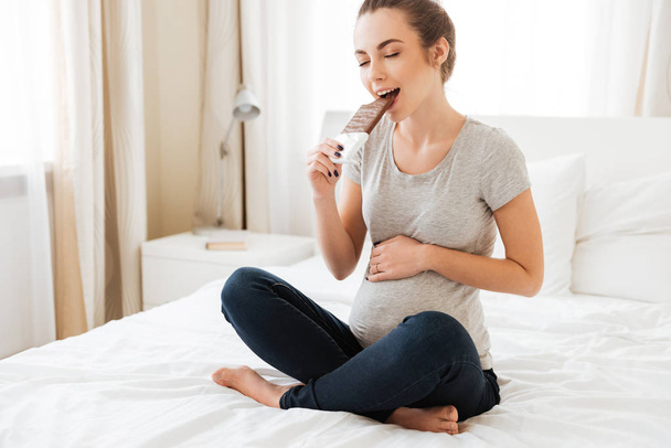 Happy pregnant young woman eating chocolate bar on bed - Foto, afbeelding