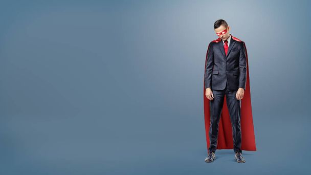 A sad businessman in a superhero red cape standing on dark blue background with his shoulders slumped. - Fotografie, Obrázek
