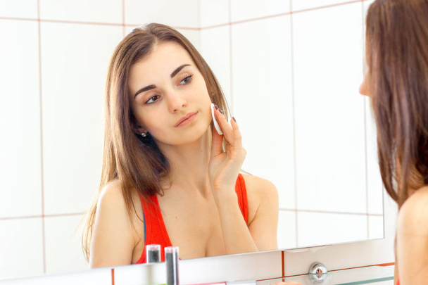 joven hermosa chica se mira en el espejo y se limpia la cara con un disco de algodón
 - Foto, Imagen