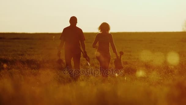 Farmers - a man and a woman walking across the field at sunset. Carry a tree seedling, a watering can and a shovel. Concept - work in the garden, volunteering, a new life - Footage, Video