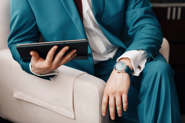El hombre de negocios tiene una tableta digital en la mano mientras está sentado en un sofá con un traje azul. reloj mecánico caro a mano con correa de cuero. camisa con gemelos
 - Foto, imagen