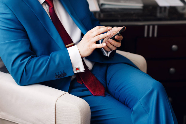 Uomo d'affari tenere un telefono in mano mentre seduto su un divano in un abito blu. a mano costoso orologio meccanico con cinturino in pelle. camicia con gemelli
 - Foto, immagini