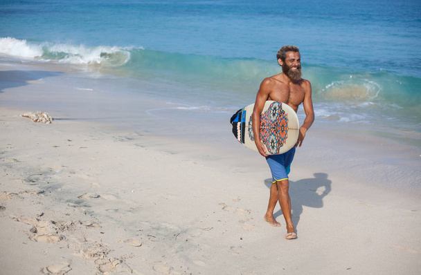 portrait of an attractive bearded curly man with a surfboard is walking on a beach, located on the right side of frame - Foto, Imagem