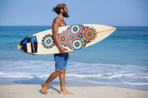 Full-length portrait of an attractive bearded curly man with a surfboard is walking on a beach - Φωτογραφία, εικόνα