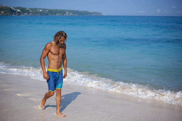 Full-length portrait of an attractive muscular bearded curly blond man with naked fit torso is walking on the beach - Foto, imagen