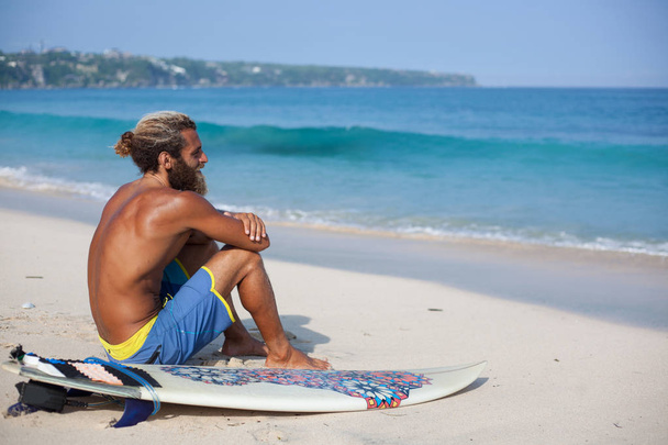 Attractive bearded curly man with a surfboard is seating with his back on a sand and looking to the ocean - Fotoğraf, Görsel
