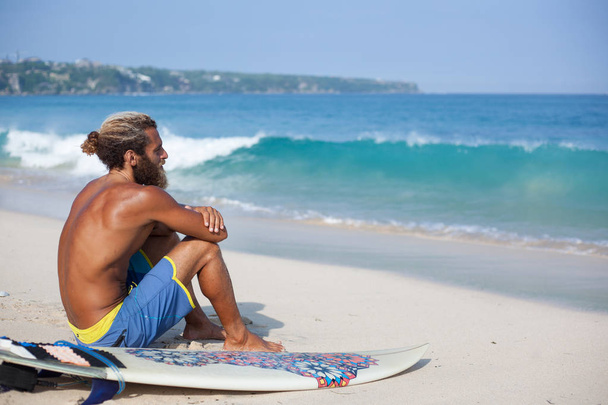 Attractive bearded curly man with a surfboard is seating with his back on a sand and looking to the ocean - Foto, immagini
