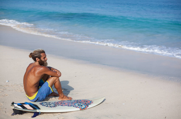 portrait of an attractive bearded curly man with a surfboard is seating with his back on a sand and looking to the ocean - Фото, зображення