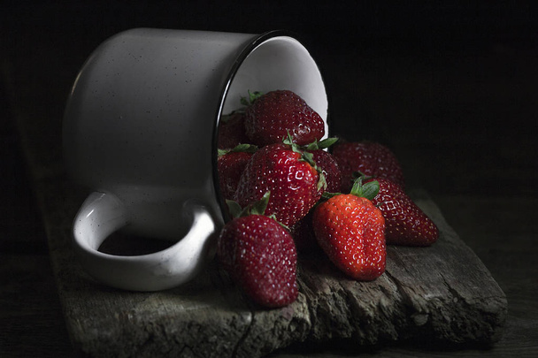 Still life with berries. Strawberries spill out of a white mug on wooden table. - Valokuva, kuva