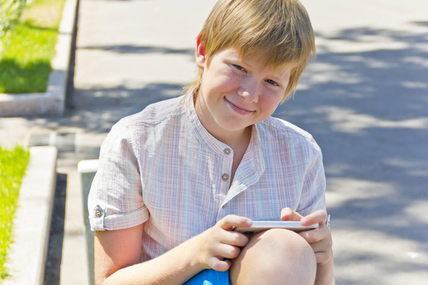 Blond boy with mobile phone sitting on a bench - 写真・画像