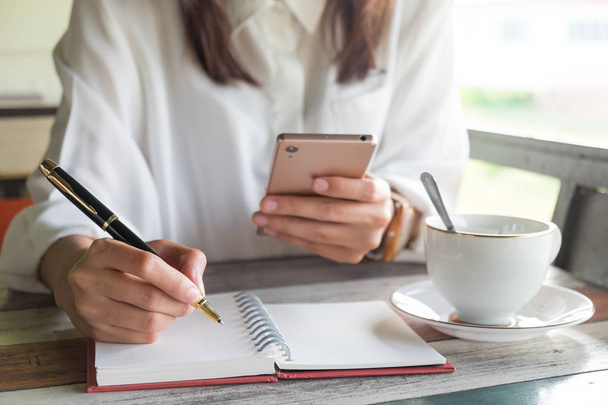 fiatal nő használ mozgatható telefon részére korlátozás valami írást üres jegyzetfüzet toll lenyomva ing felszerelni, és kávé csésze mellé üzembe. Ez a kép a bushiness, az oktatás és a portré-fogalom - Fotó, kép