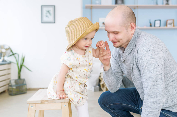 Bonito pai jovem ajuda a filhinha a se levantar de uma cadeira
 - Foto, Imagem