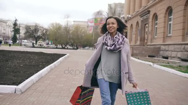 Dolly shot of Attractive mixed race girl dancing and have fun while walking down the street with bags. Happy young woman walking after shopping on mall sale - Πλάνα, βίντεο
