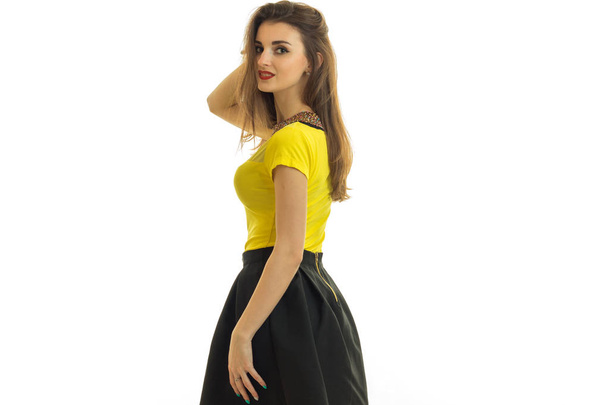 a charming young girl in a skirt and yellow shirt stands turning sideways and smiling - Fotó, kép