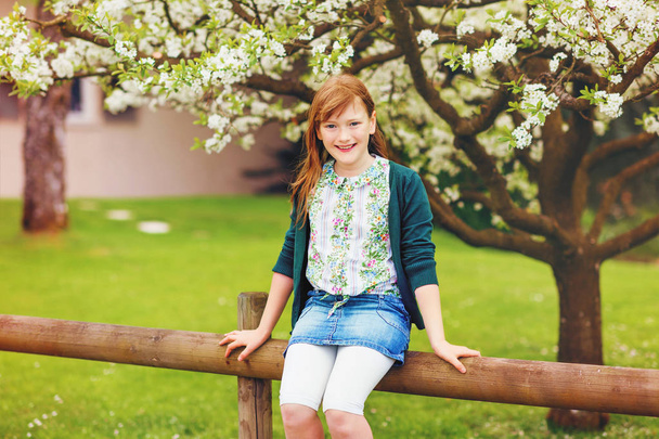 Outdoor spring portrait of happy young kid girl sitting on the fence, wearing denim skirt and green jacket - Photo, Image