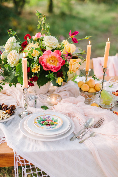 picnic, comida, verano, concepto de vacaciones - primer plano en parte de la mesa festivamente decorada con un hermoso ramo de rosas y peonías, velas en candelabros, albaricoques, cerezas, pera, cristalería
. - Foto, Imagen