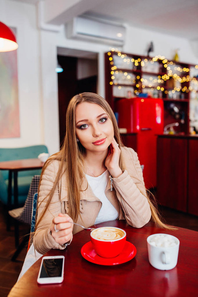 Beautiful young woman with a red cup of coffee at a cafe. Woman drinking hot latte coffee at cozy coffee shop - Fotoğraf, Görsel