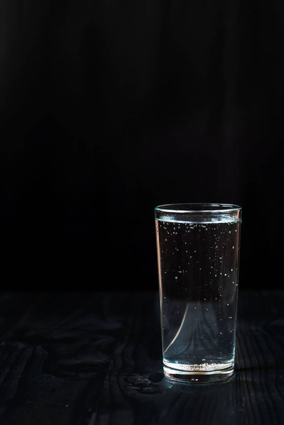 Μόλις ένα ποτήρι νερό σε μαύρο φόντο. Μεταλλικό νερό σε ένα ποτήρι σε ένα σκοτεινό ξύλινο τραπέζι. - Φωτογραφία, εικόνα