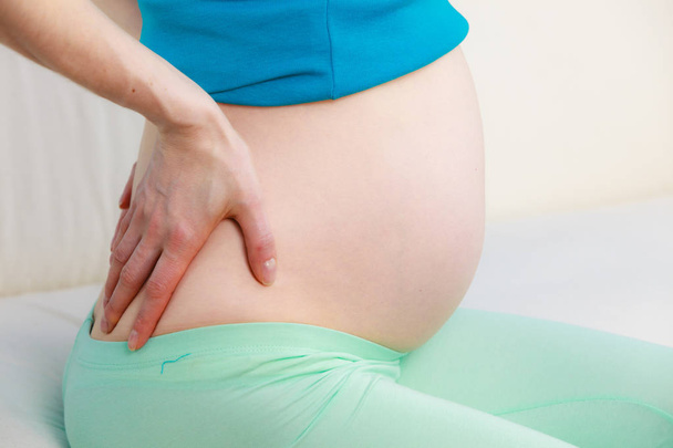 Mulher grávida sentada no sofá segura a mão em suas costas
 - Foto, Imagem