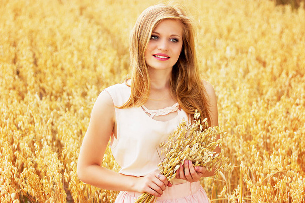 Краса сільської місцевості. Портрет привабливої молодої рудої волохатої дівчини, одягненої в білу сукню, що тримає пшеницю в руках, стоячи на жовтому пшеничному полі
. - Фото, зображення