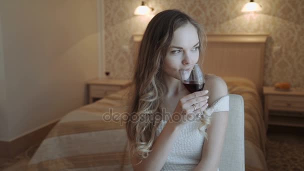 Young business woman relaxing at her hotel bedroom Drinks from a glass of red wine at the window. in luxurious room interior. Young beautiful girl in white dress - Felvétel, videó
