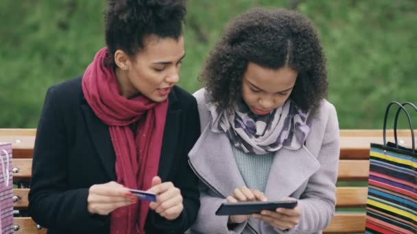 Two attractive mixed race women with paper bags sitting on bench and using tablet computer and credit card for online shopping - Séquence, vidéo